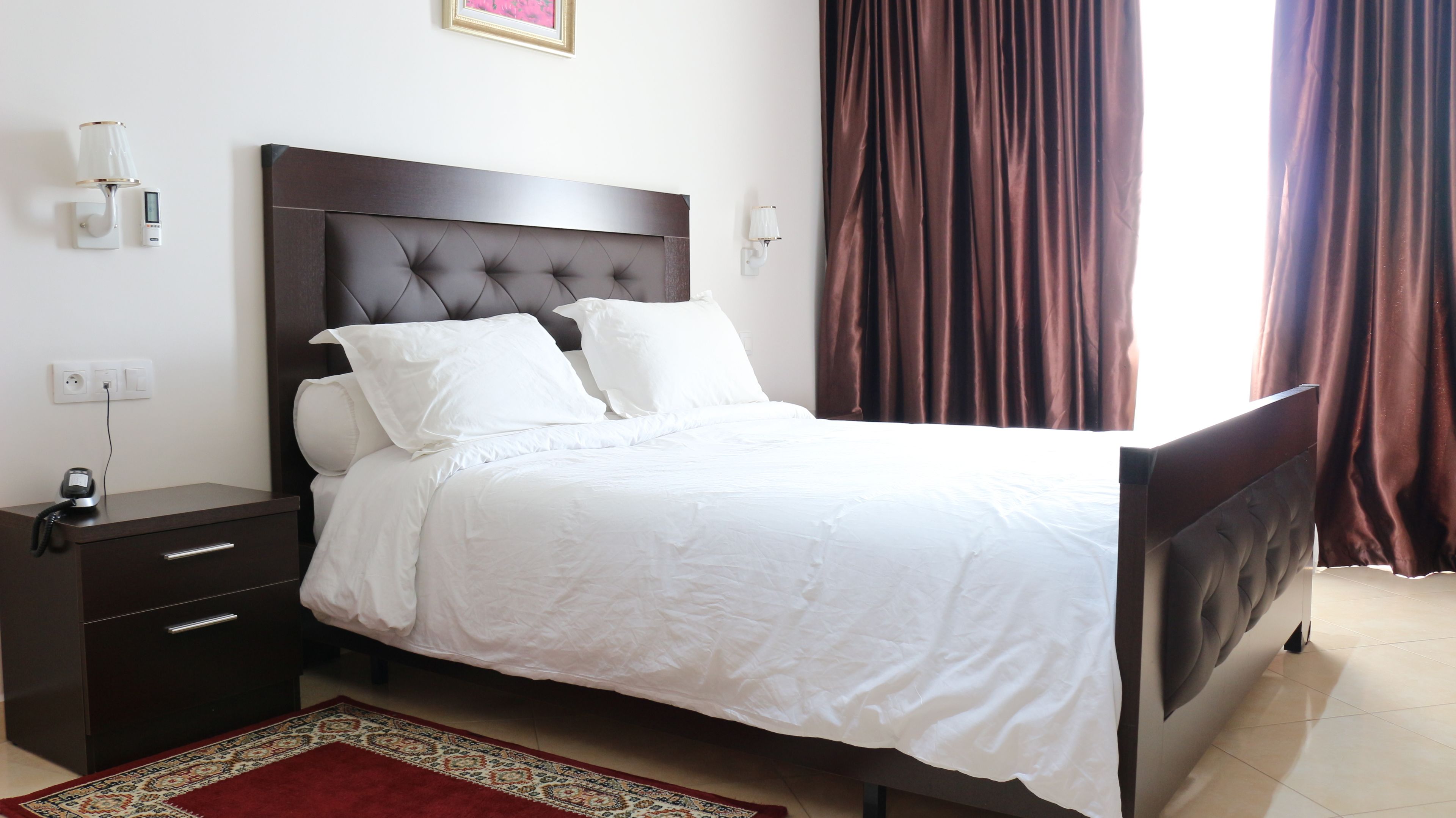 A neatly made bed with white linens, a dark headboard, and a bedside table with a lamp and a phone.