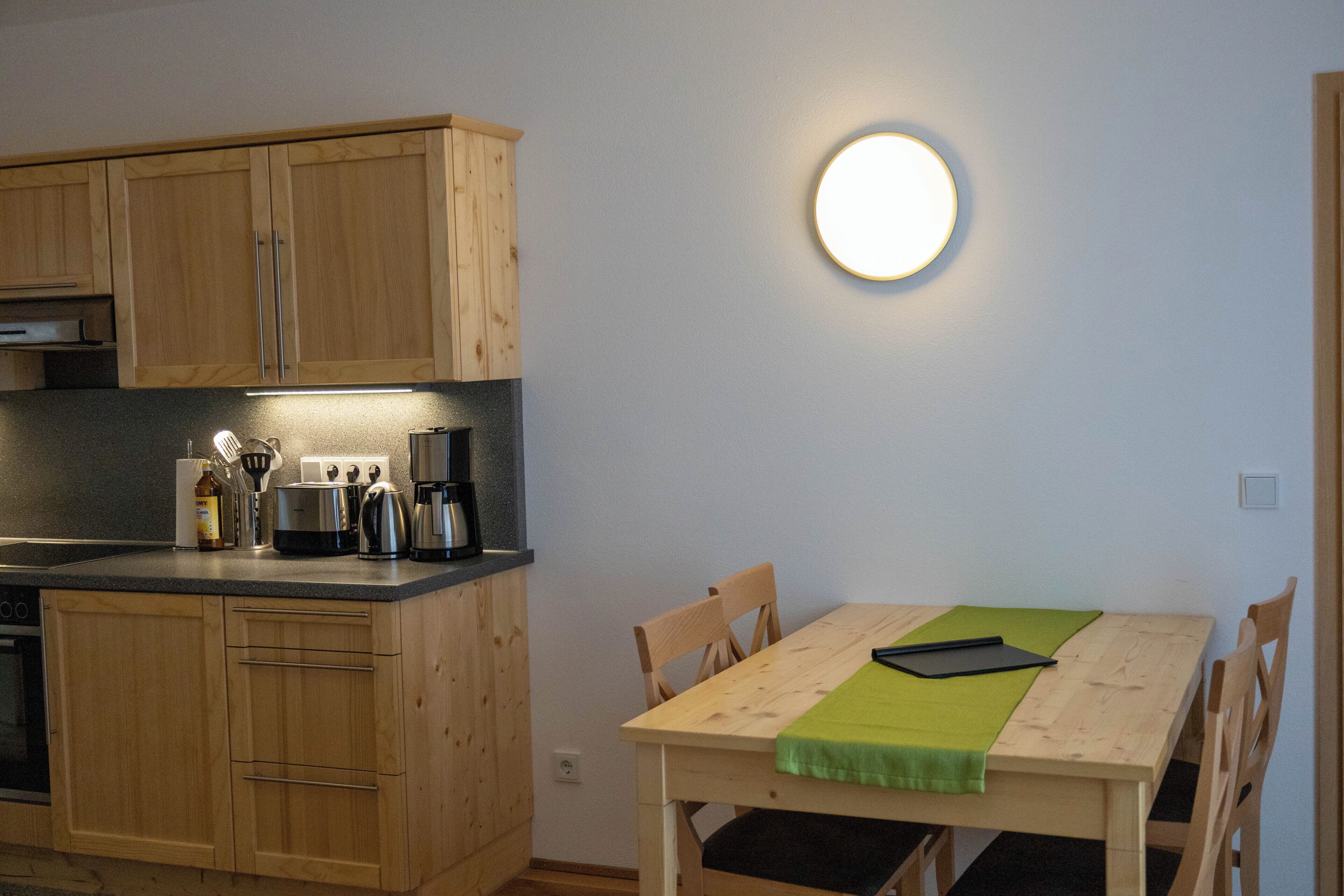 Een keuken met houten kastjes, een aanrecht met apparatuur, een eettafel met stoelen en een wandlamp.