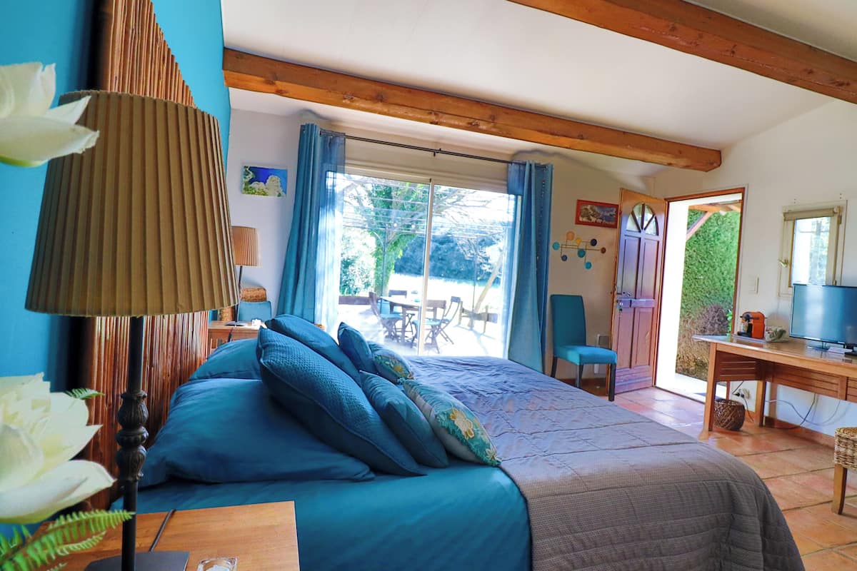 A bedroom with a bed, a desk, and a chair. There is a window with curtains, a lamp, and a plant.