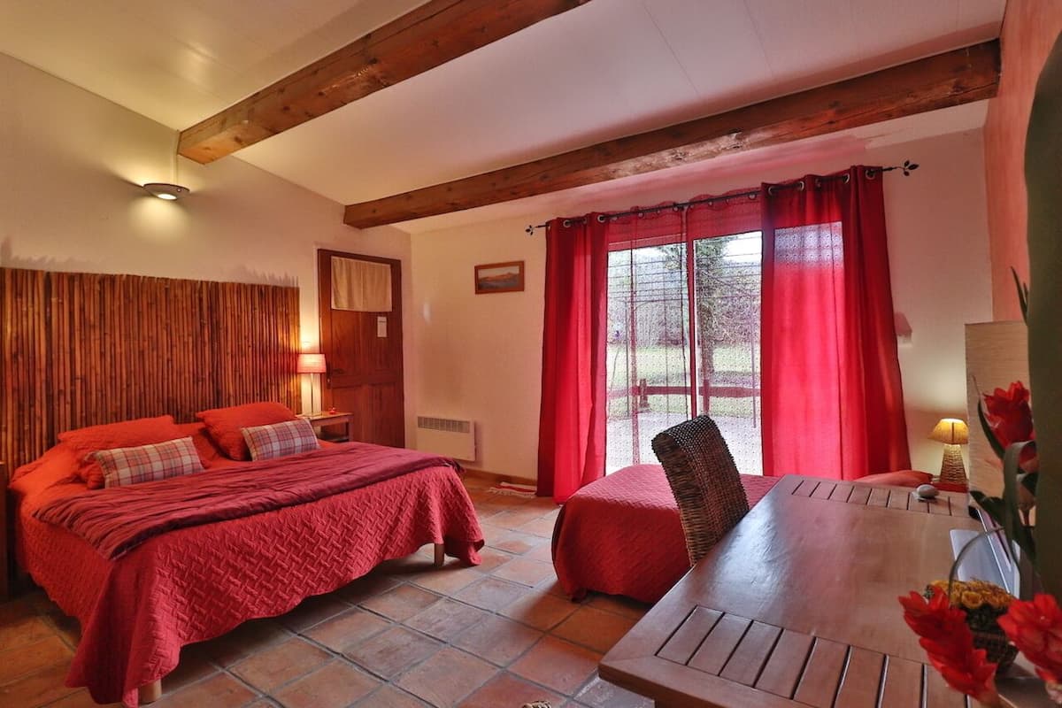 A bedroom with a wooden ceiling, a bed with red bedding, a table with a chair, and a window with red curtains.