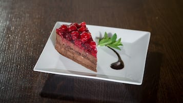 A slice of chocolate cake with red fruit topping on a white plate, accompanied by a mint leaf and chocolate drizzle.