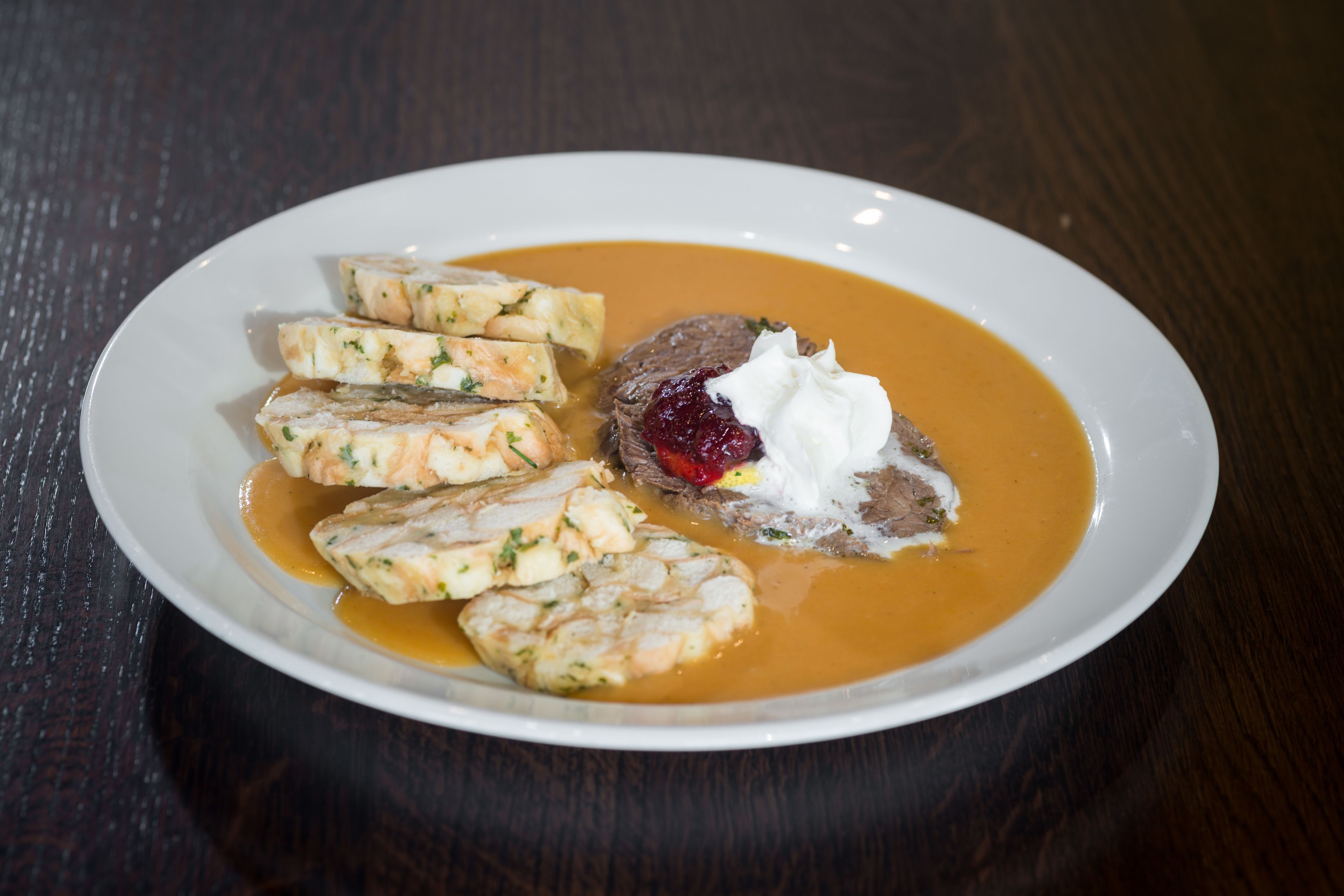 En hvid tallerken med cremet suppe, skiver af kød og en klat fløde på et mørkt træbord.