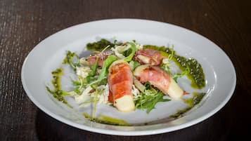 Ein weißer Teller mit einem Salat bestehend aus Blattsalaten, Käse und Prosciutto auf einem dunklen Holztisch.