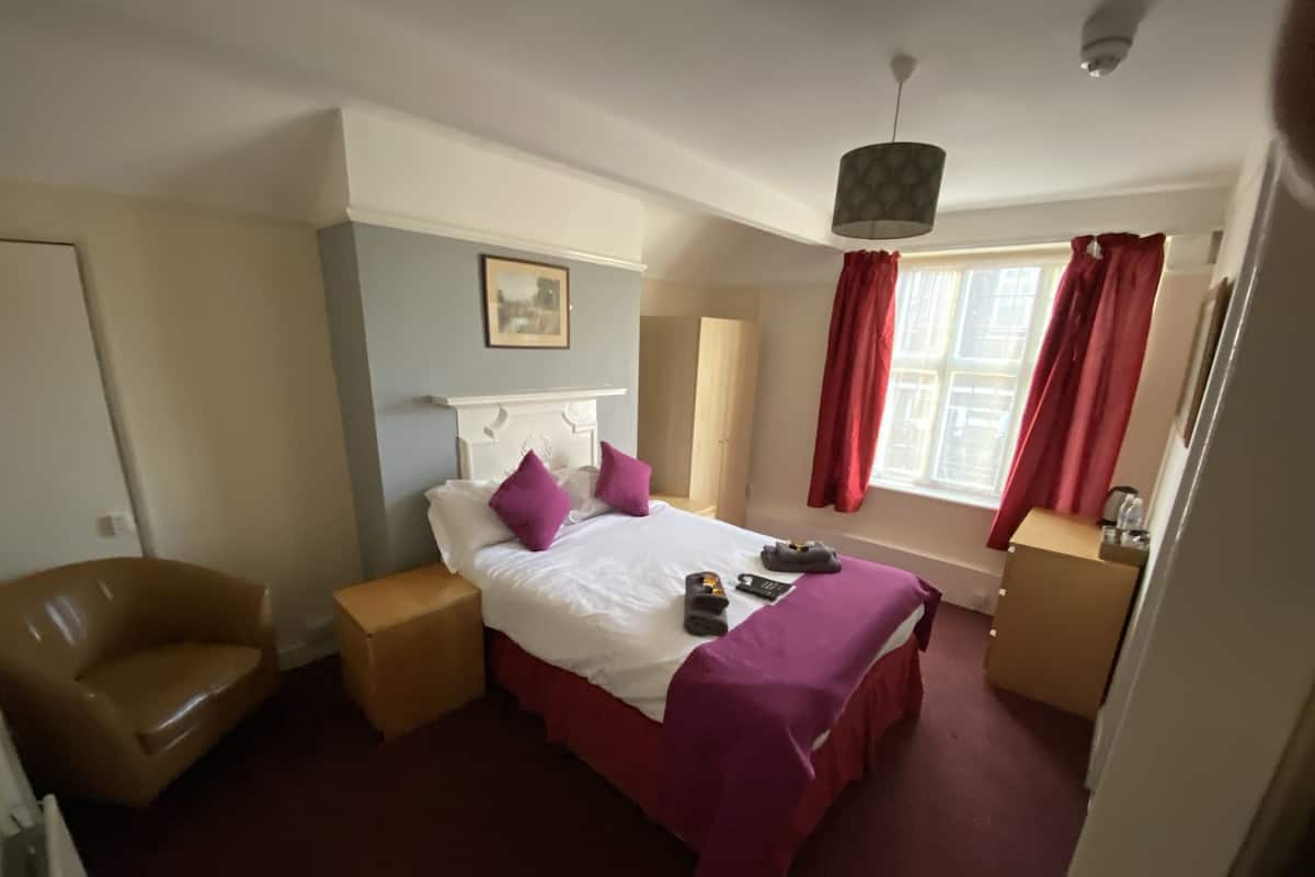 Habitación de hotel con cama, una silla, mesita de noche, ventana con cortinas y una lámpara en el techo.