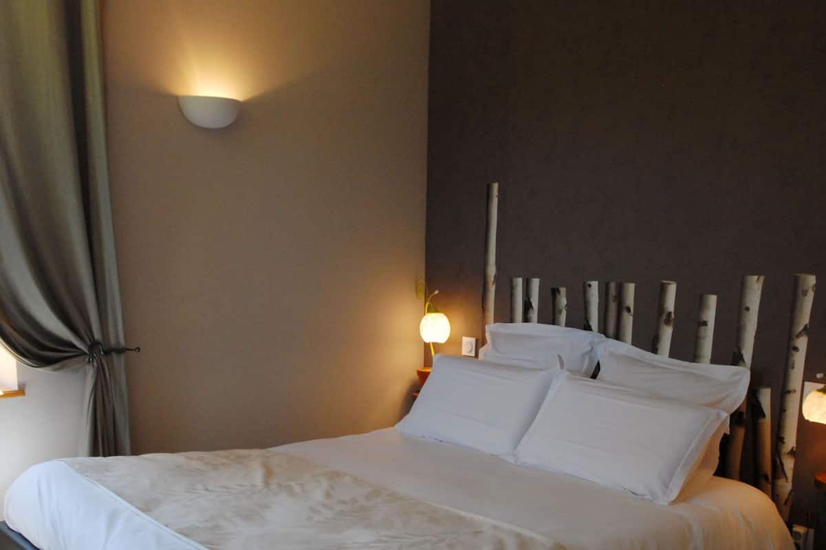 Una cama bien hecha con sábanas blancas, un aplique de pared y un ventanal con cortinas.