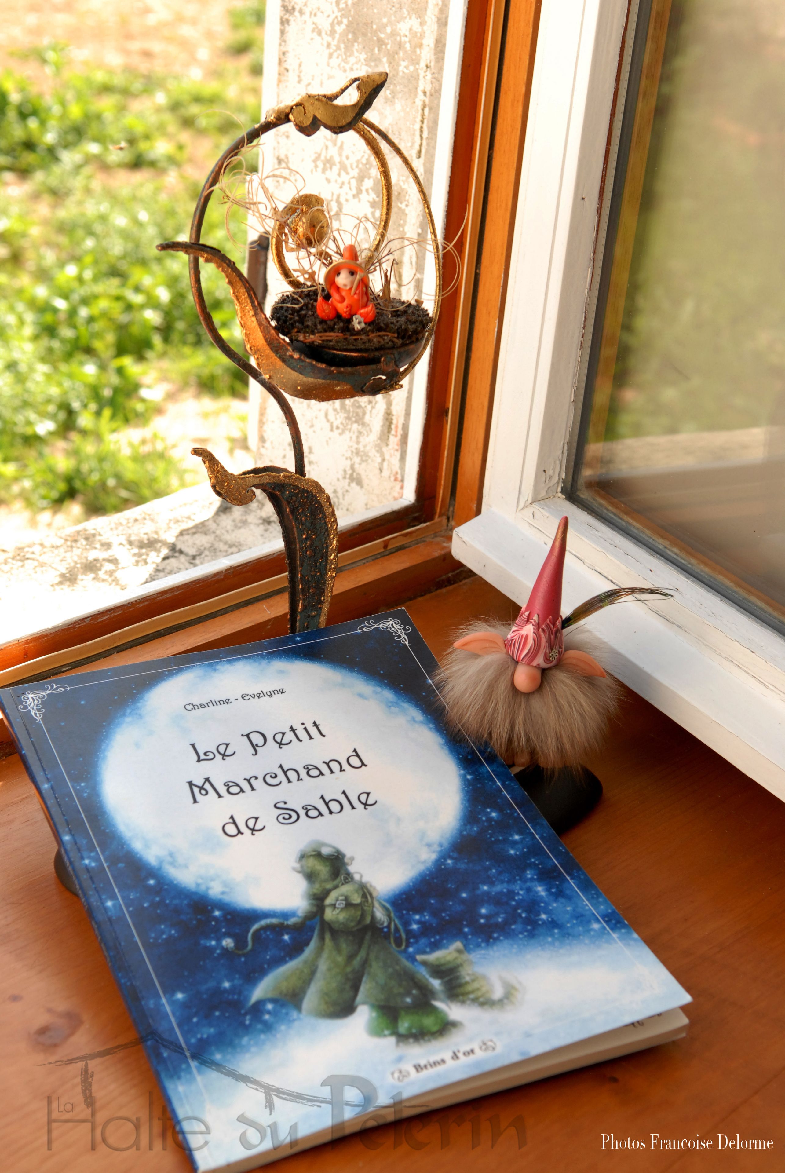 Un livre intitulé « Le Petit Marchand de Sable » est posé sur une surface en bois, près d’une fenêtre, et un présentoir en métal décoratif se trouve à proximité.