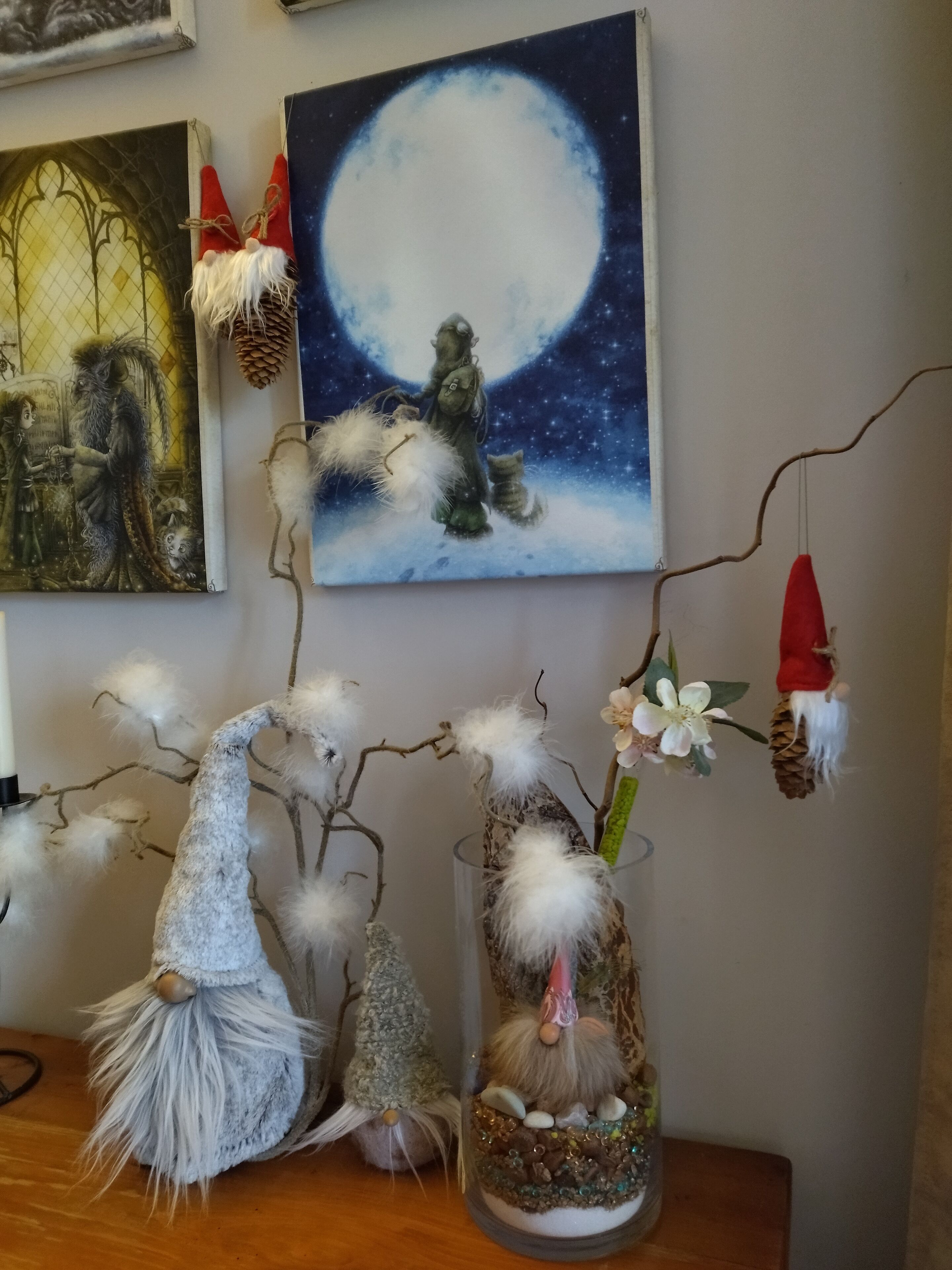 A room with a large painting of a moonlit scene, a decorative tree branch with hanging ornaments, and a glass vase with a gnome figurine.