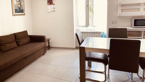 A living room with a brown sofa, a dining table with chairs, a window with blinds, and a wall-mounted TV. - Appartamenti St'Andrea (Levanto)
