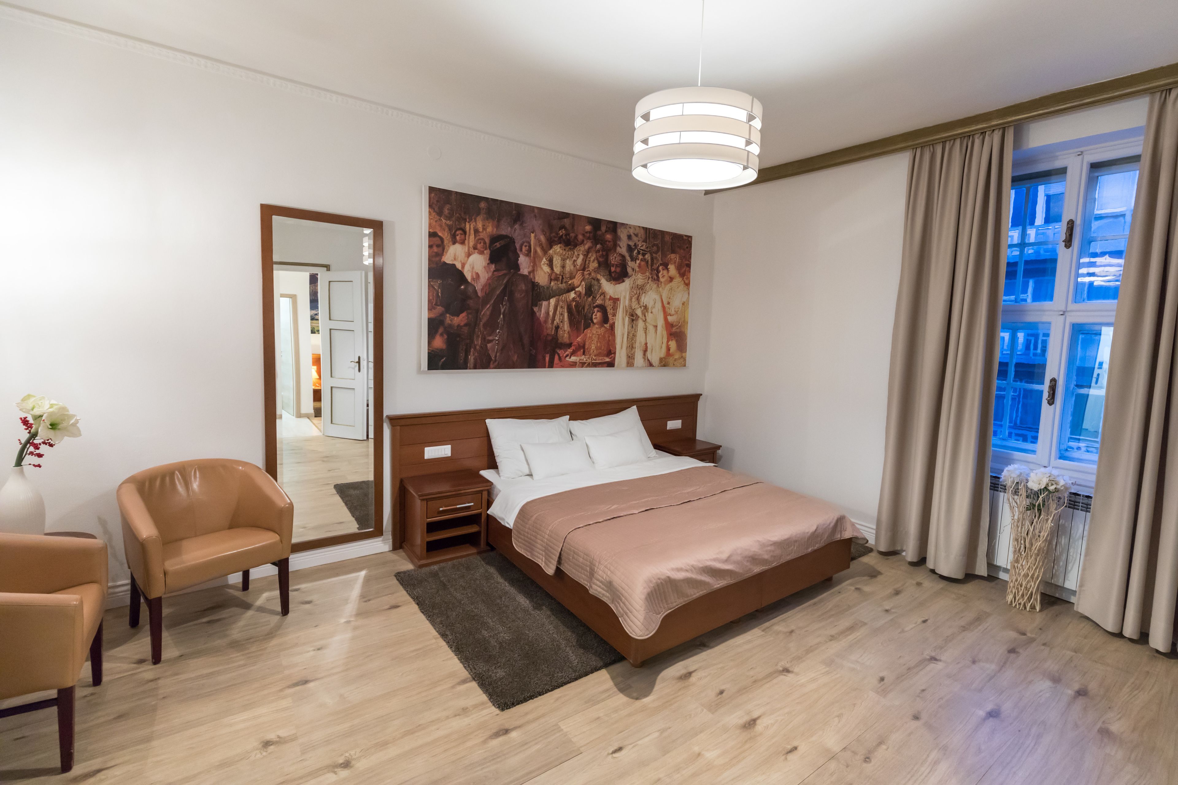 Een slaapkamer met een houten bed, een nachtkastje, een stoel, een groot schilderij aan de muur en een raam met gordijnen.