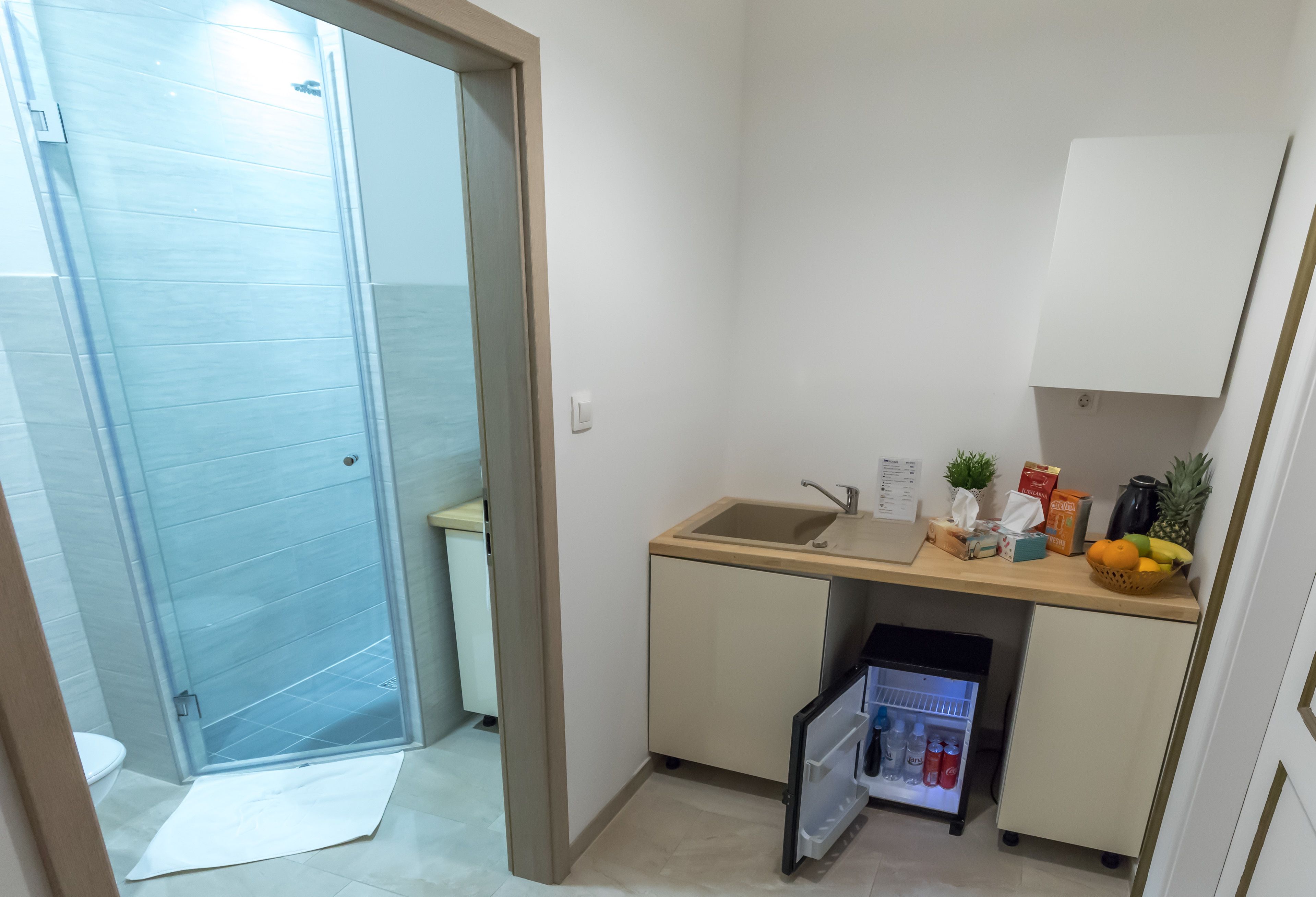A compact bathroom with a glass shower door, a sink with a small countertop, and a mini fridge stocked with beverages.