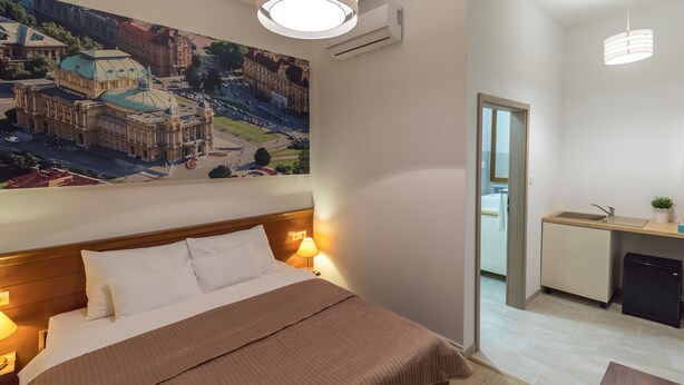 A hotel room with a large bed, a painting of a building, a small kitchenette, and a door leading to another room. - Bacu Apartments Zagreb (Zagreb)