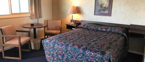 Habitación de hotel con cama, dos sillas, una mesita, una lámpara y un cuadro en la pared.