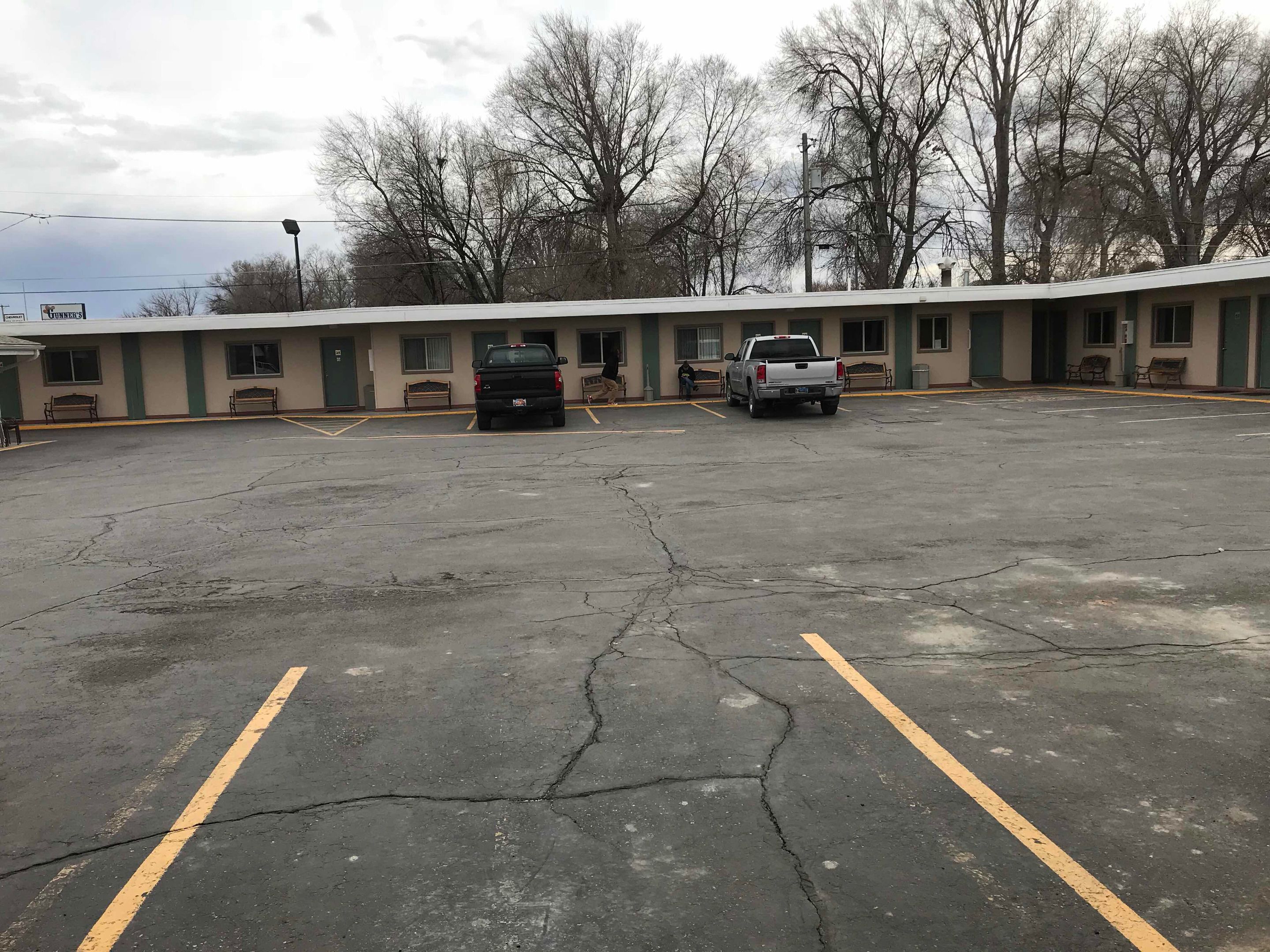 Un estacionamiento con dos vehículos, un edificio con varias habitaciones y una fila de árboles al fondo.