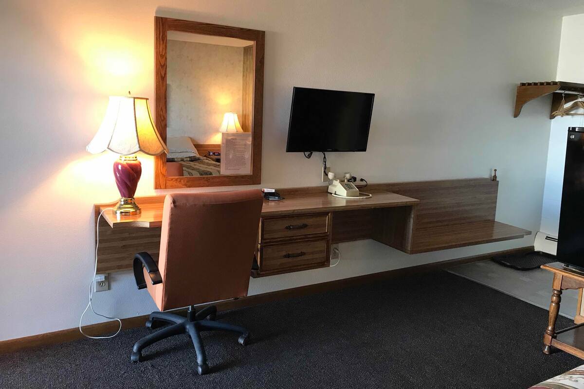 A hotel room with a wooden desk, a chair, a television, a lamp, and a mirror.
