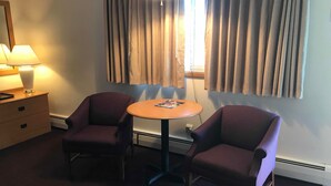 Une chambre d’hôtel avec deux fauteuils violets, une petite table ronde, une lampe, une table de chevet et une fenêtre avec des rideaux.