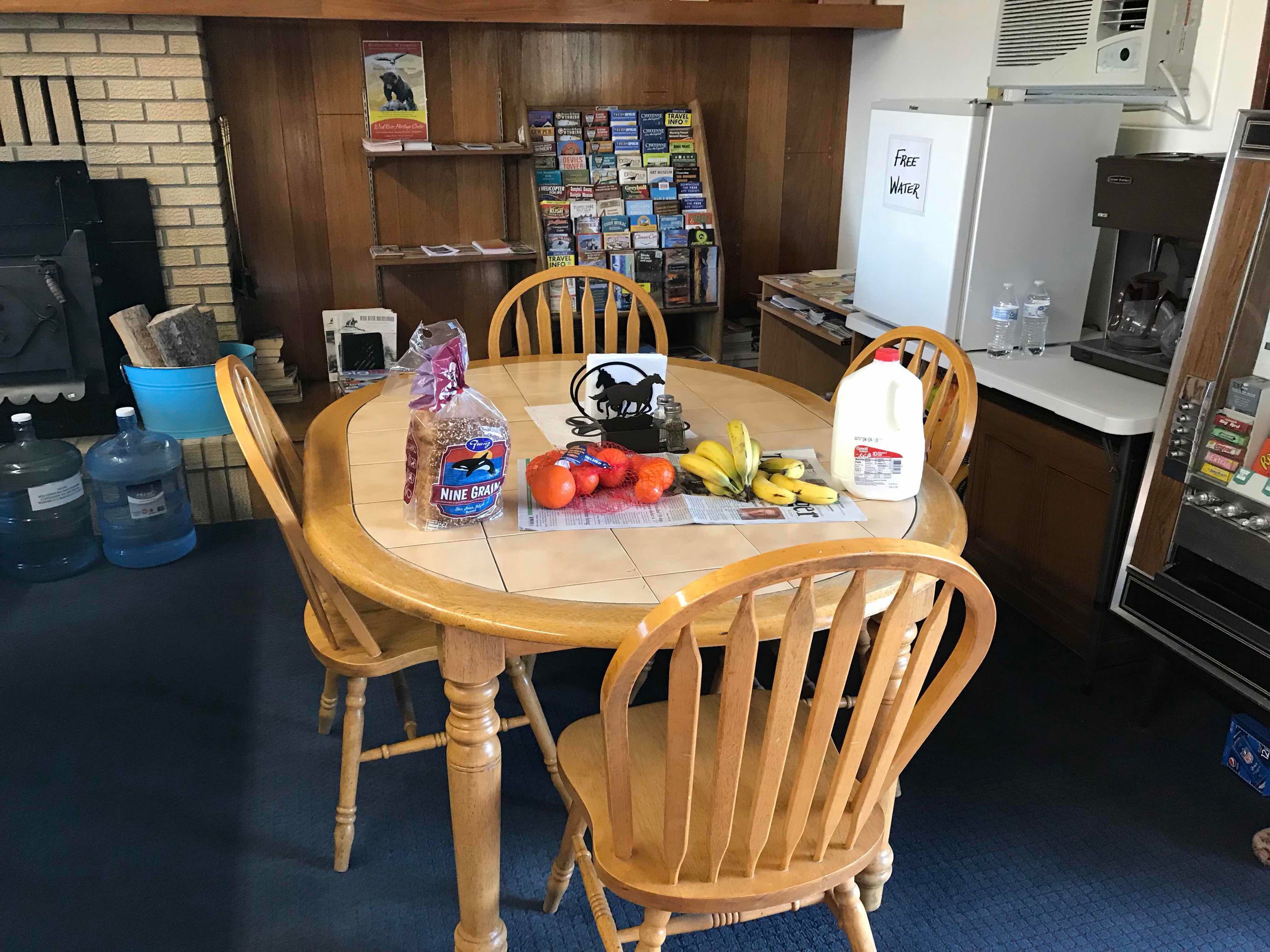 A dining area with a round wooden table, chairs, and a variety of food items including bread, oranges, bananas, and a milk jug. There are water dispensers and a refrigerator in the background.