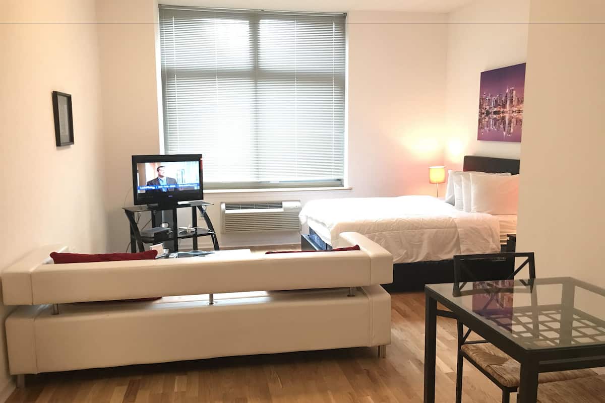Een hotelkamer met een bed, een televisie op een standaard, een bank, een tafel en een raam met jaloezieën.