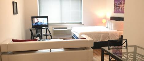 Habitación de hotel con cama, televisor en un mueble, sofá, mesa y ventana con persianas.