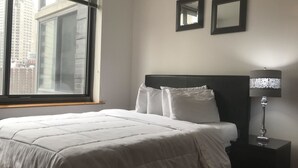 A hotel room with a large bed, a nightstand with a lamp, two wall mirrors, and a window with blinds. - Downtown Luxury Suites (Jersey City)