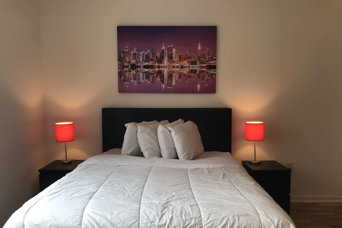 Een net opgemaakt bed met een witte dekbedovertrek, twee nachtkastjes met rode lampen, en een stadslandschap schilderij aan de muur.