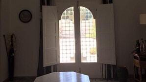 Une pièce avec une table blanche, des chaises, un luminaire suspendu et une fenêtre dont les volets sont ouverts.