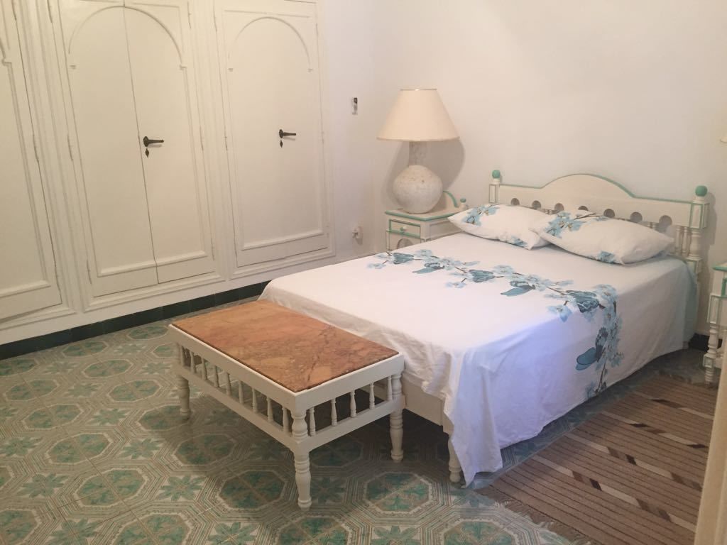 A bedroom with a bed, a bedside table with a lamp, a wooden bench, and patterned floor tiles.