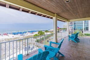 A covered balcony with blue Adirondack chairs, overlooking a beach with umbrellas and a pool.