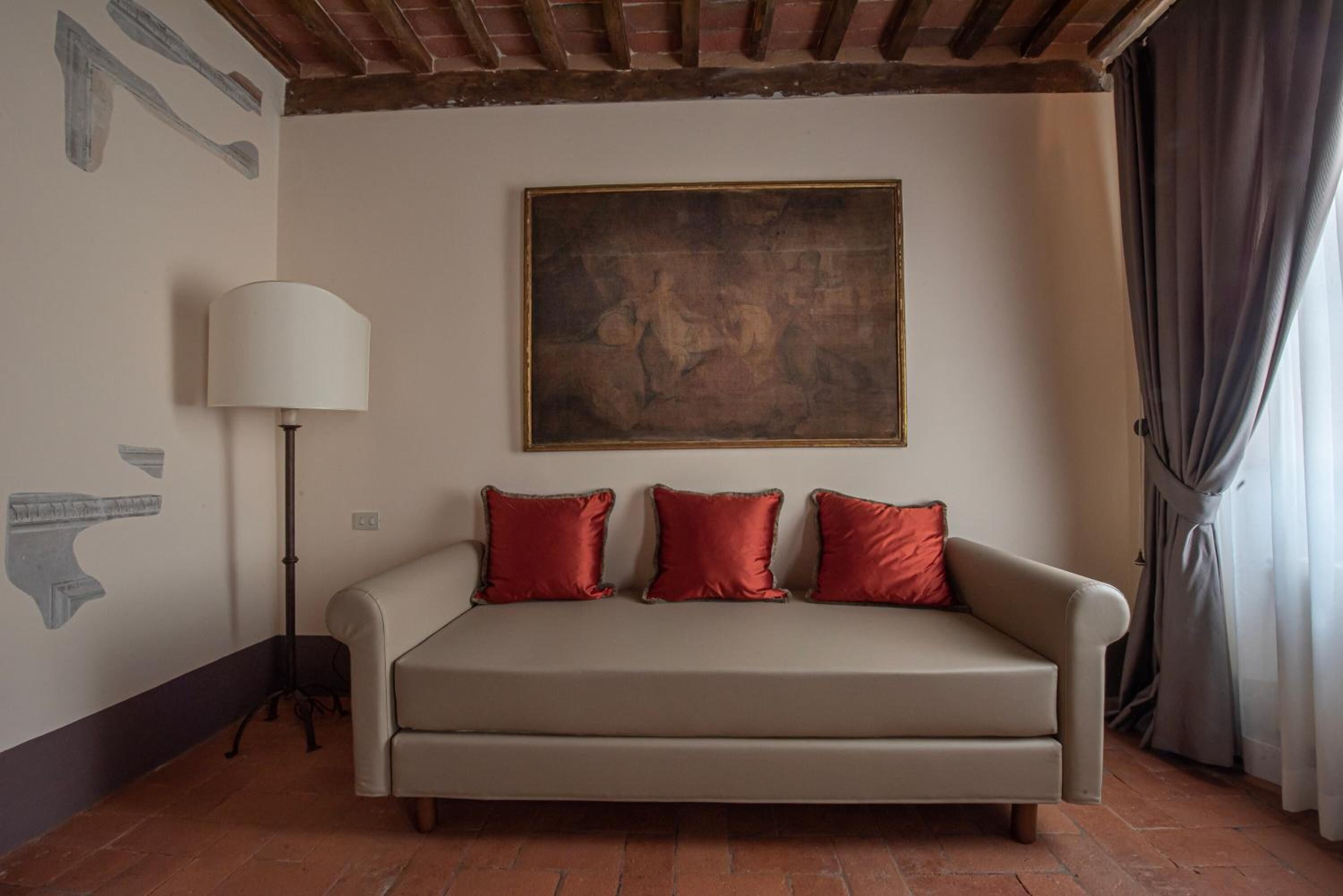Una habitación con un sofá color beige, tres almohadas rojas, una lámpara de pie y un cuadro grande en la pared.