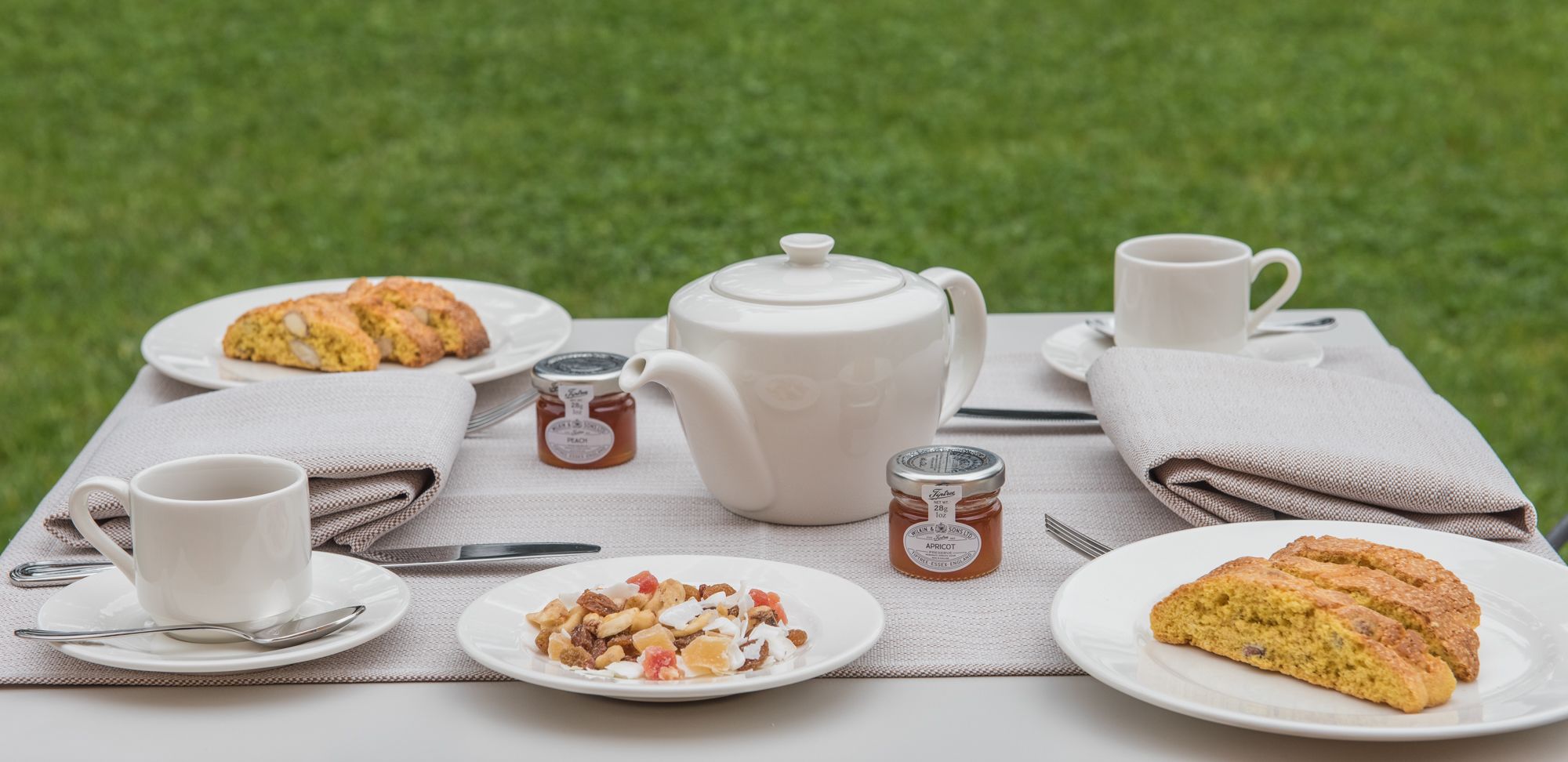 A picnic setup with a white teapot, two cups, a plate of scones, a plate of nuts, and two jars of jam on a tablecloth.