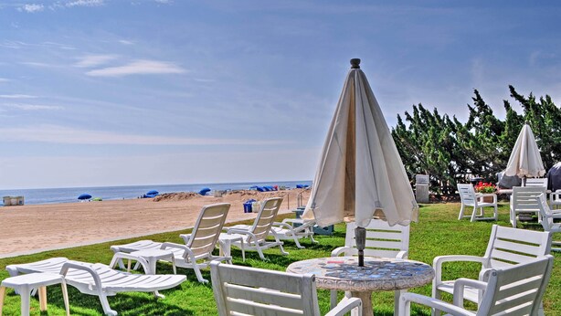 Outdoor seating with white plastic chairs and umbrellas, a round table, and a view of the beach. - Colorful Virginia Beach Studio - Steps to Beach! (Virginia Beach)