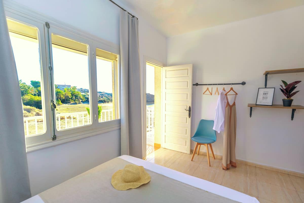 Une chambre à coucher avec un lit, une vue sur l’extérieur, une chaise et une étagère ornée d’une plante et d’une citation encadrée.