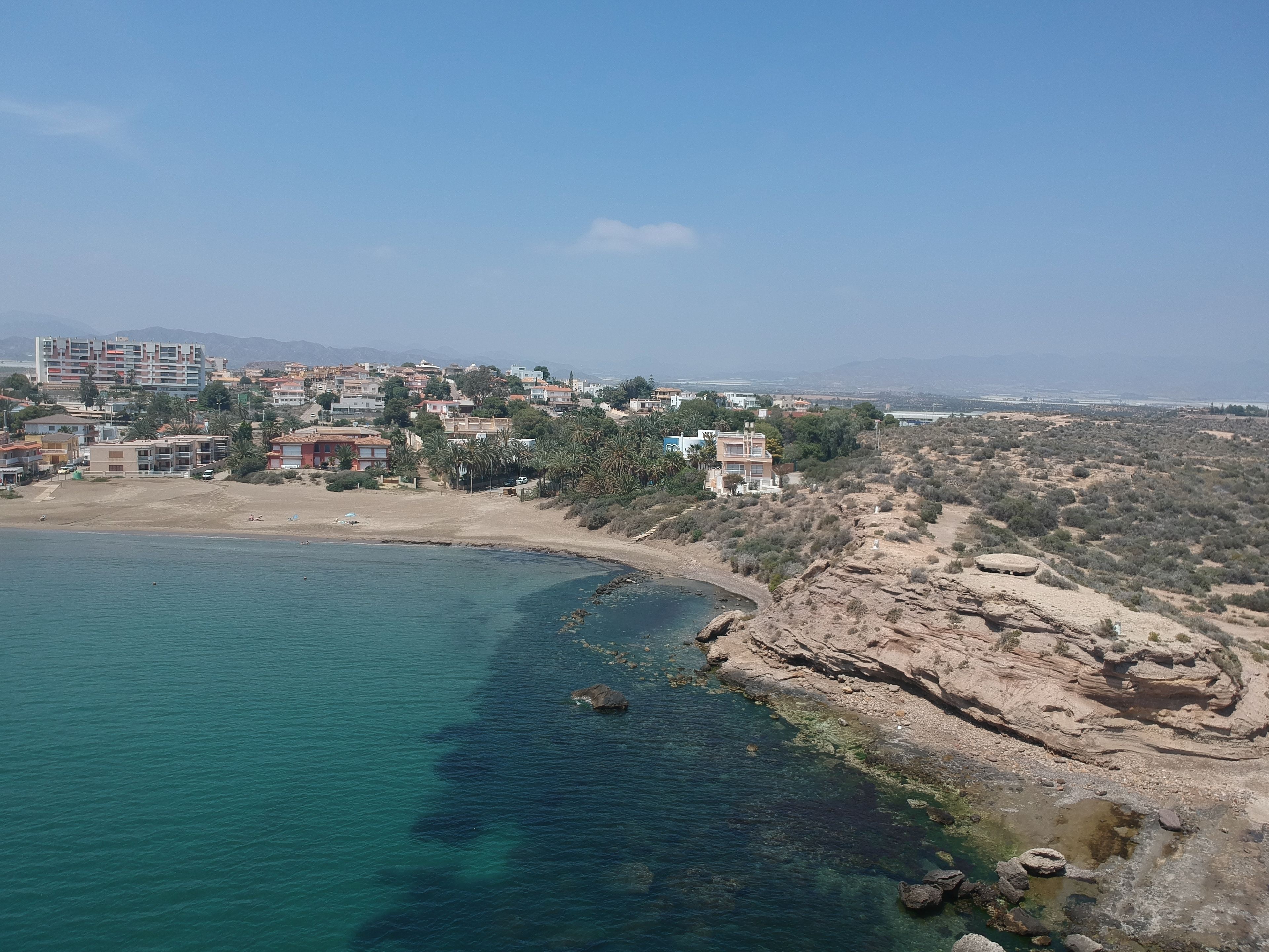 Un pueblo costero con edificios a lo largo de la orilla, aguas azules cristalinas y terreno rocoso.