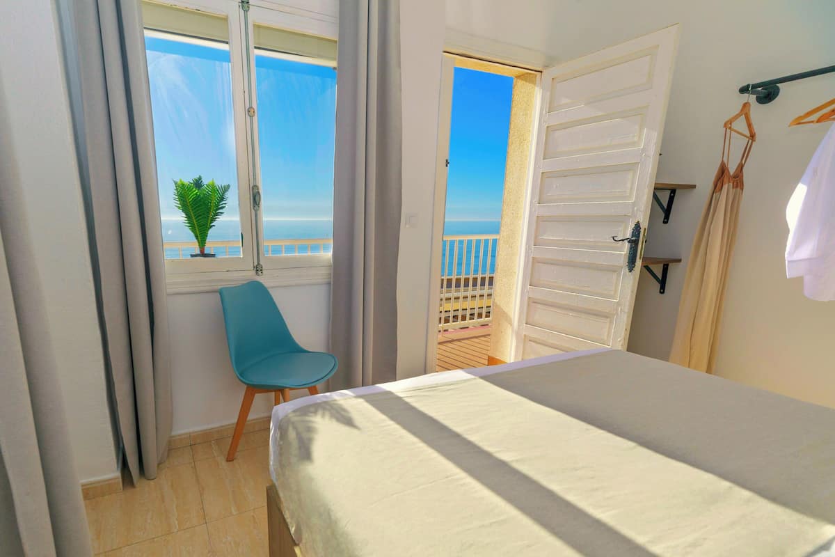 Une chambre à coucher comprenant un lit, une chaise, une fenêtre donnant sur la mer et une porte menant à un balcon.