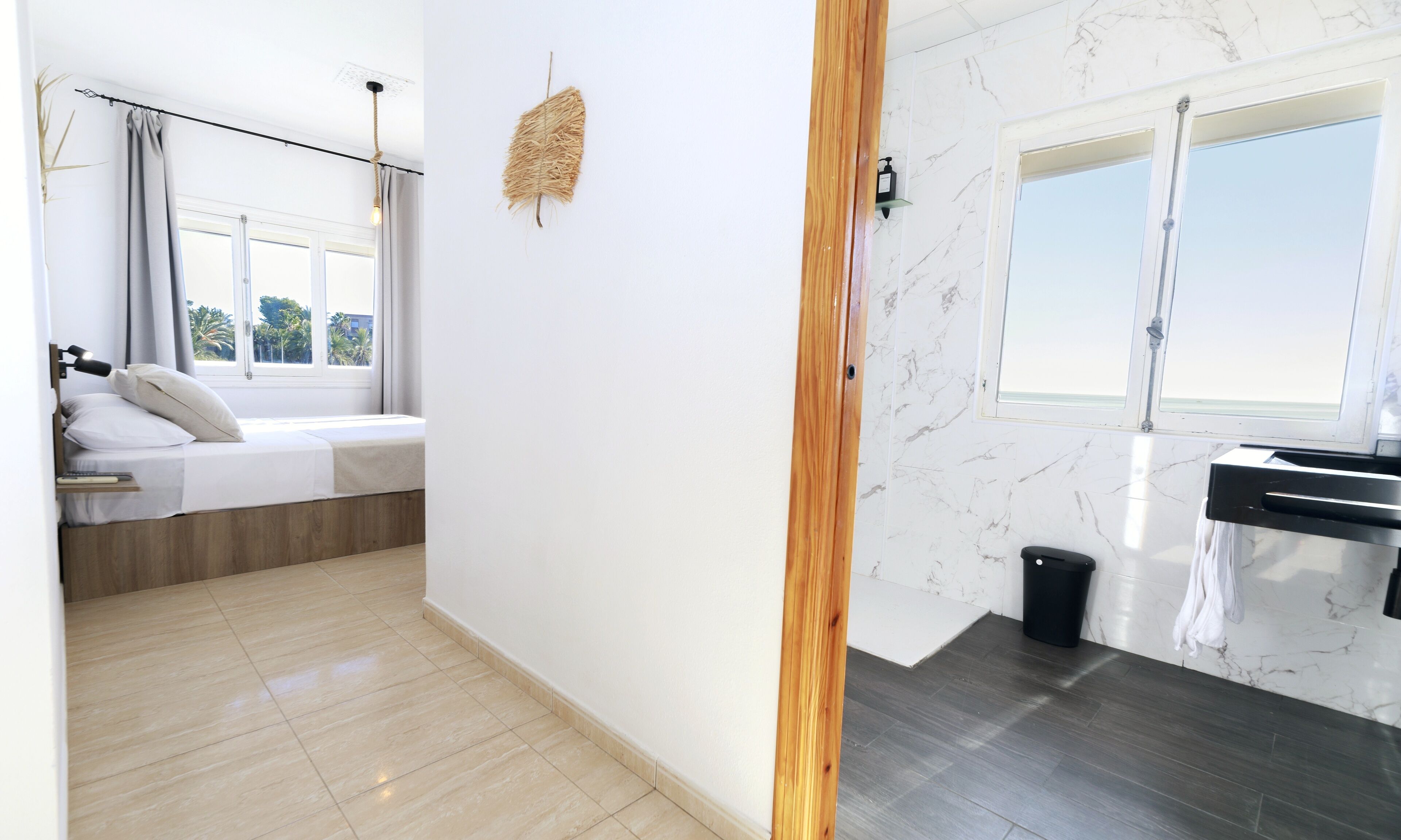 Une chambre d’hôtel moderne comprenant un lit, une petite table de chevet, une fenêtre avec des rideaux, un luminaire fixé au mur et une salle de bain avec un lavabo et un miroir.