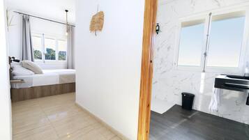 Quarto de hotel moderno com cama, mesinha de cabeceira, janela com cortinas, luminária na parede e banheiro com pia e espelho.