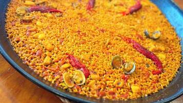 Una paellera grande y poco profunda llena de un plato amarillo, posiblemente un tipo de arroz o comida a base de granos, decorada con chiles rojos y almejas.