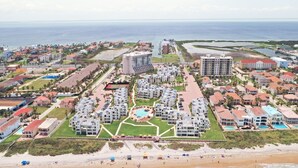 An aerial view of a coastal resort with multiple buildings, a central pool area, and a beach in the foreground.