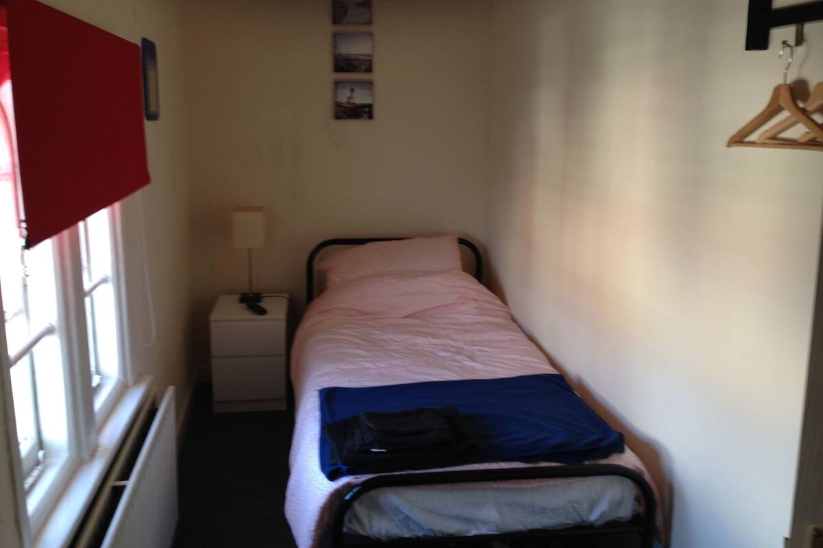 Ein schmales Zimmer mit einem Einzelbett, einem Nachttisch mit Lampe, einem roten Vorhang und einem hölzernen Kleiderbügel.