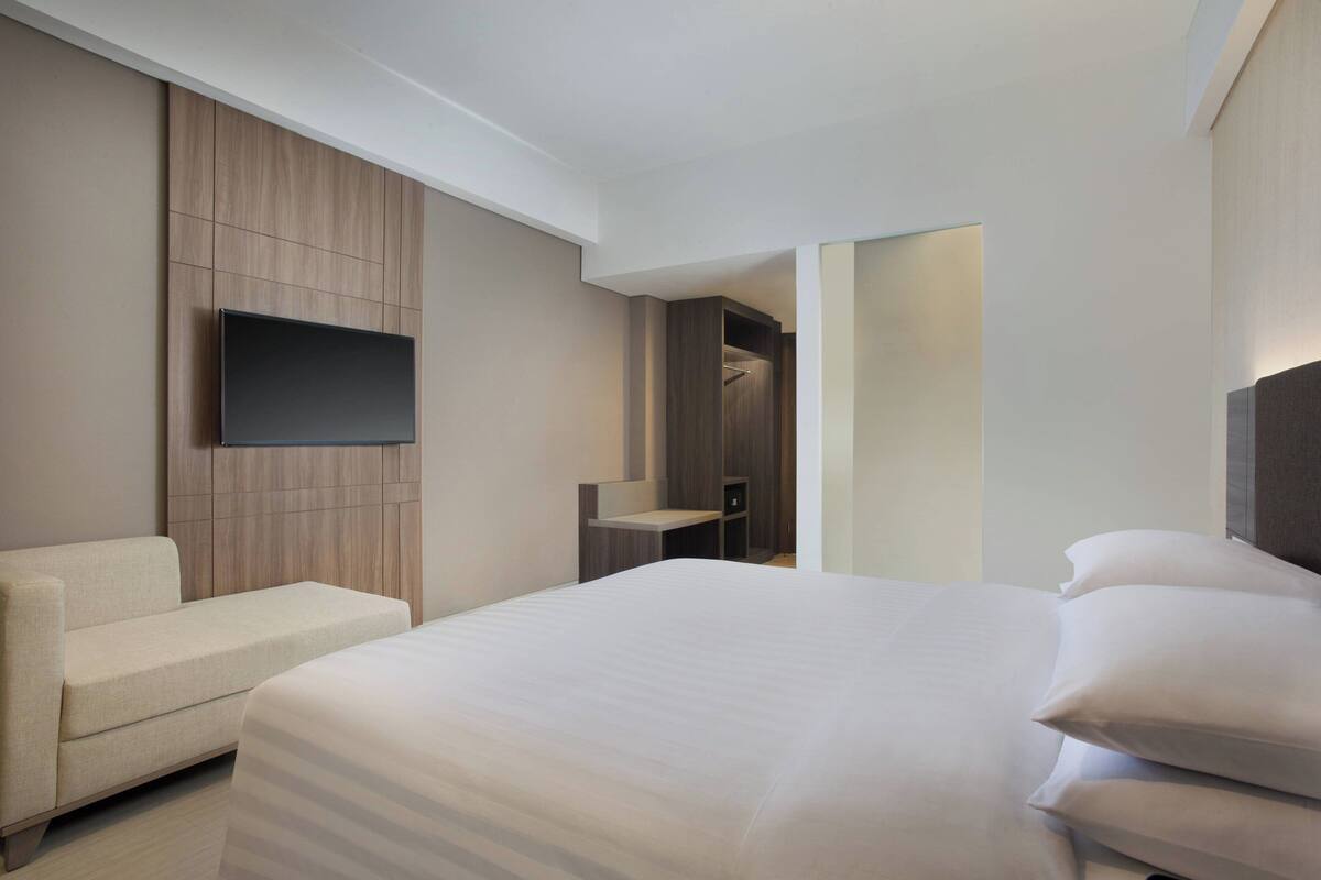 A modern hotel room with a large bed, a flat-screen TV mounted on the wall, a seating area, and a walk-in closet.