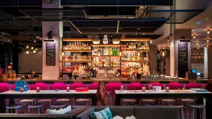 Zona de bar con varias estanterías llenas de botellas, un espacio con taburetes y una barra con caja registradora.