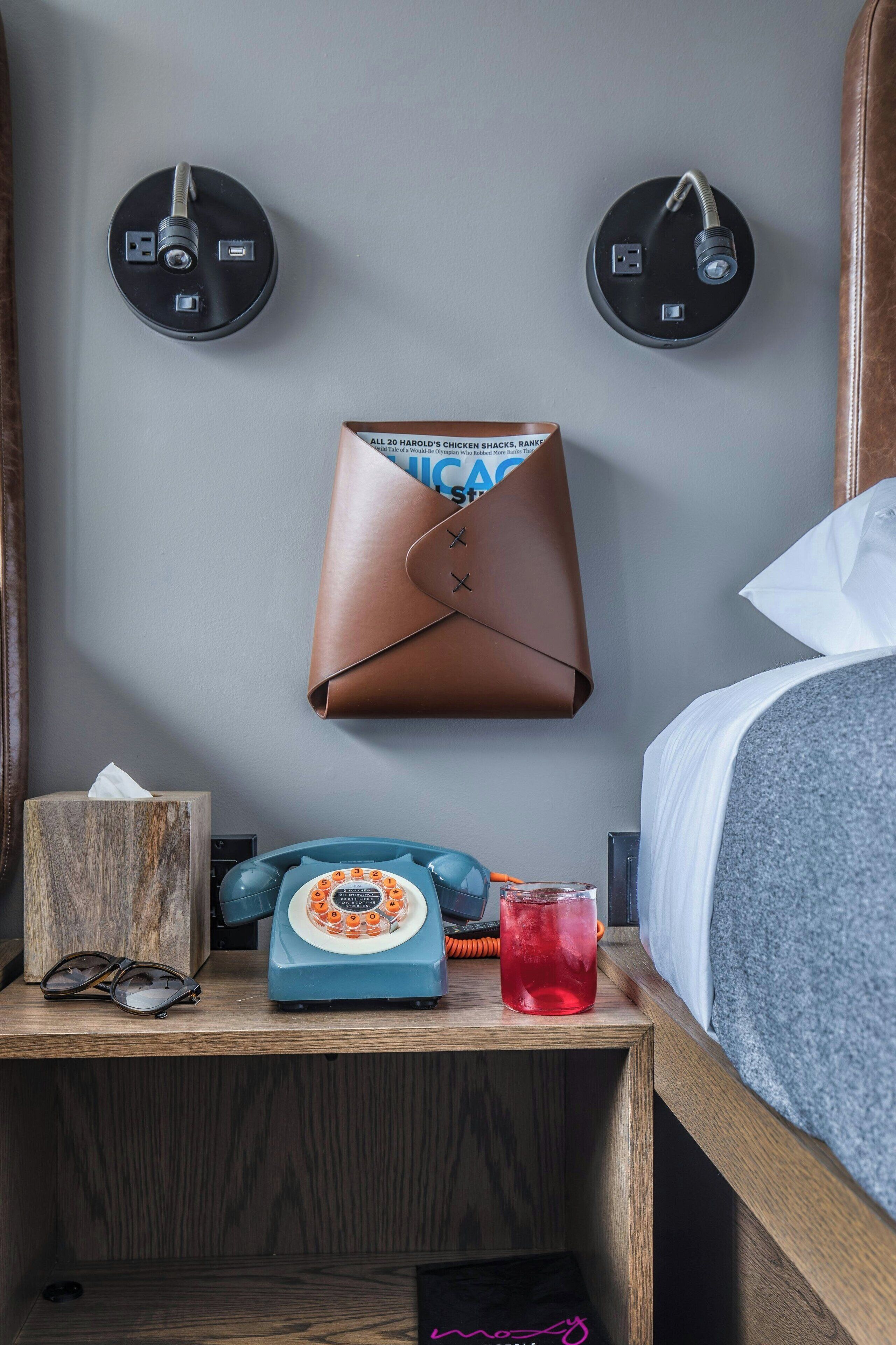 Ein Hotelzimmer mit einem hölzernen Nachttisch, einem altmodischen blauen Wählscheibentelefon, einer Papiertuchbox und einer braunen Lederhandtasche, die an der Wand hängt.