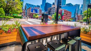 Un área de estar al aire libre con una mesa de madera y taburetes de metal, con vista a un paisaje urbano con edificios modernos y una calle.