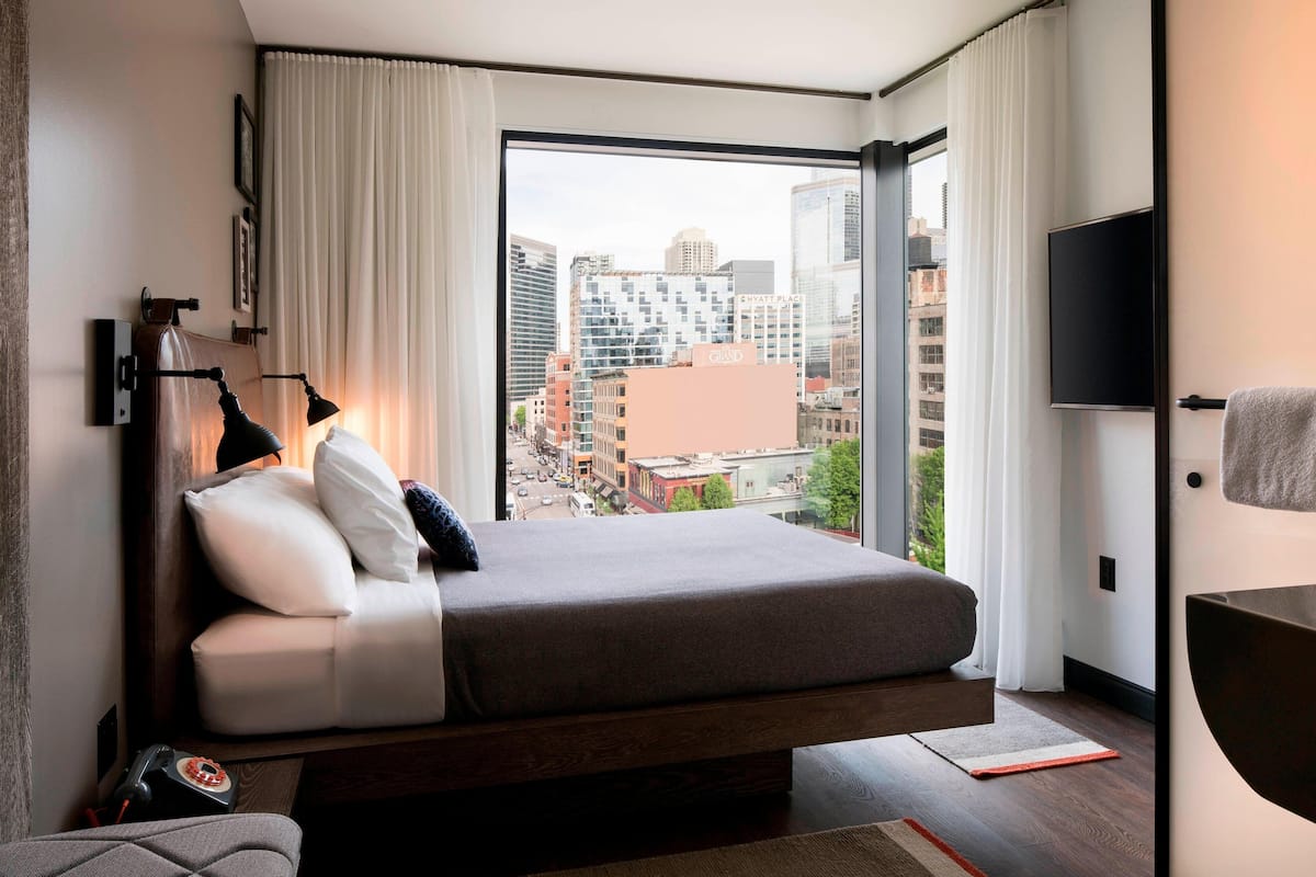Habitación de hotel moderna con una cama grande, vistas de la ciudad, un televisor de pantalla plana y un escritorio con lámpara.