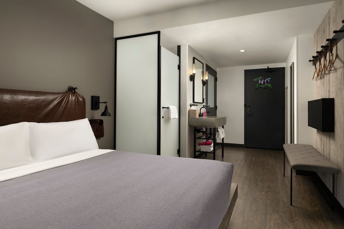 Una habitación de hotel moderna con una cama grande, un banco, una tocador con espejo y una puerta con un letrero de neón.