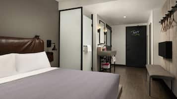 A modern hotel room with a large bed, a bench, a vanity with a mirror, and a door with a neon sign.