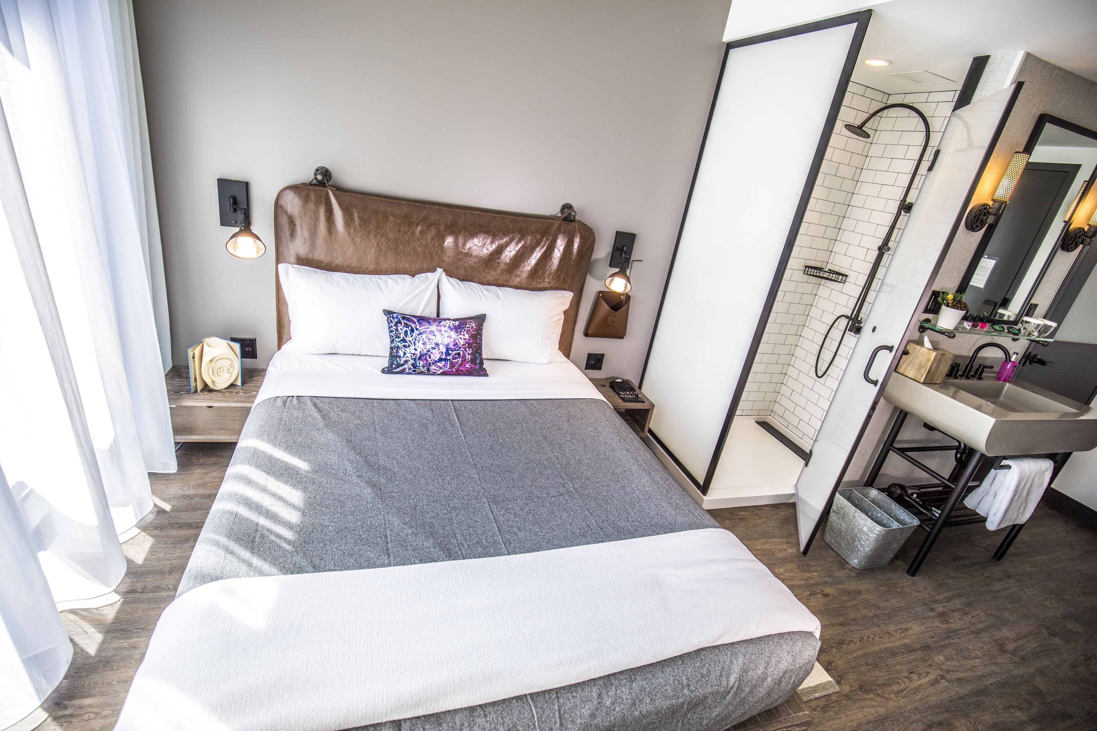A modern hotel room with a large bed, a shower, a vanity with a mirror, and a bedside table with a lamp.
