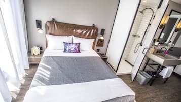 A modern hotel room with a large bed, a shower, a vanity with a mirror, and a bedside table with a lamp.