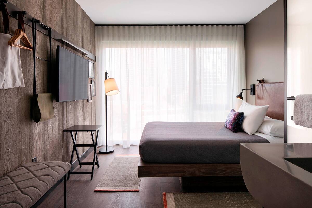 Un dormitorio moderno con cama, mesita de noche, banqueta y televisión montada en la pared.