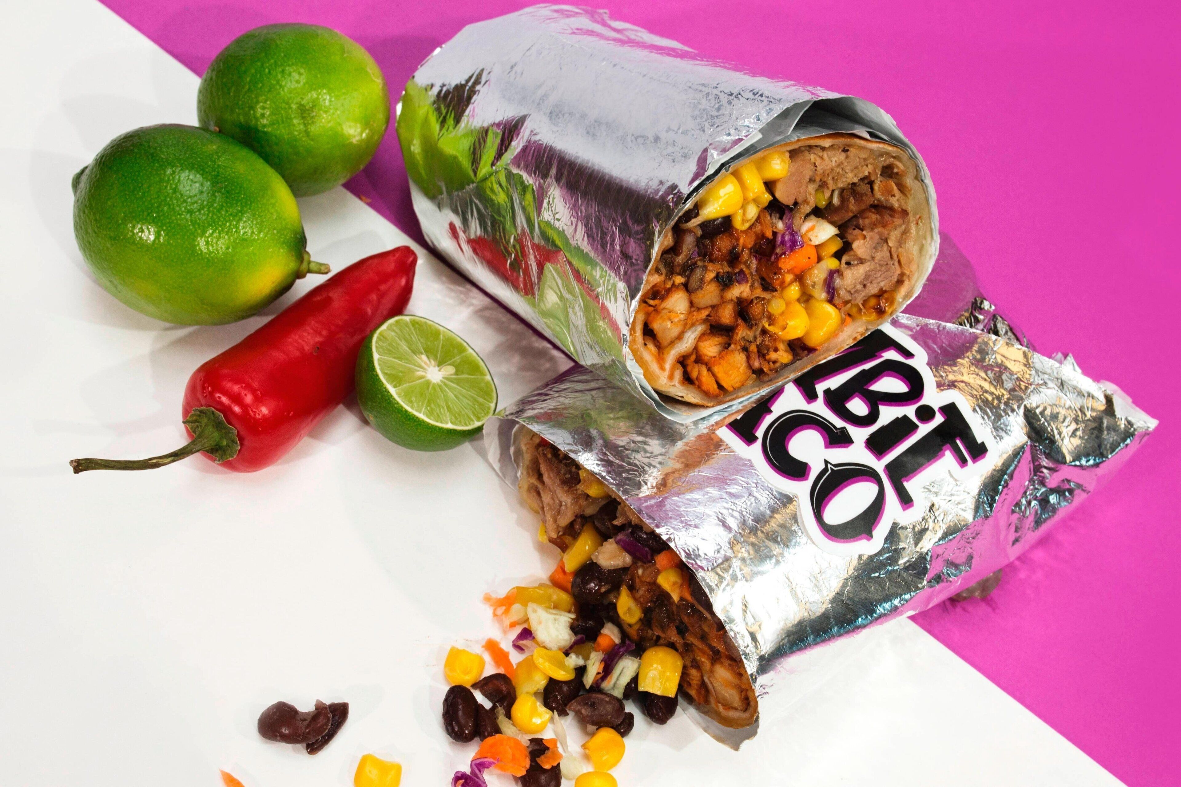 A burrito with visible ingredients like beans, corn, and meat, accompanied by lime, a red chili pepper, and two limes on a white surface.