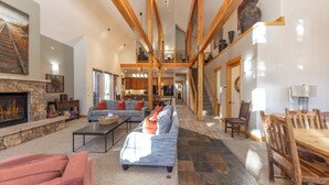 A spacious living area with a fireplace, a sofa, a coffee table, and a dining table. There is a staircase leading to an upper level. - Sawmill Run Lodge: Pet-Friendly, Walk to Peak 8, Hot Tub, Grill, Shuttle (Breckenridge)